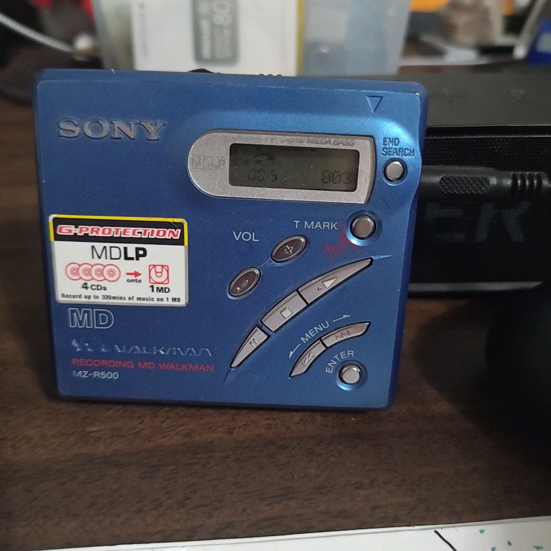 Blue Sony MZ-R500 recording MiniDisc Walkman standing on a desk, showing its display, control buttons and connected cable.