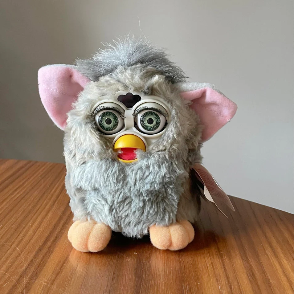 Grey-and-white Furby toy with large green eyes, pink ears, a yellow beak and peach-coloured feet, standing on a wooden table.