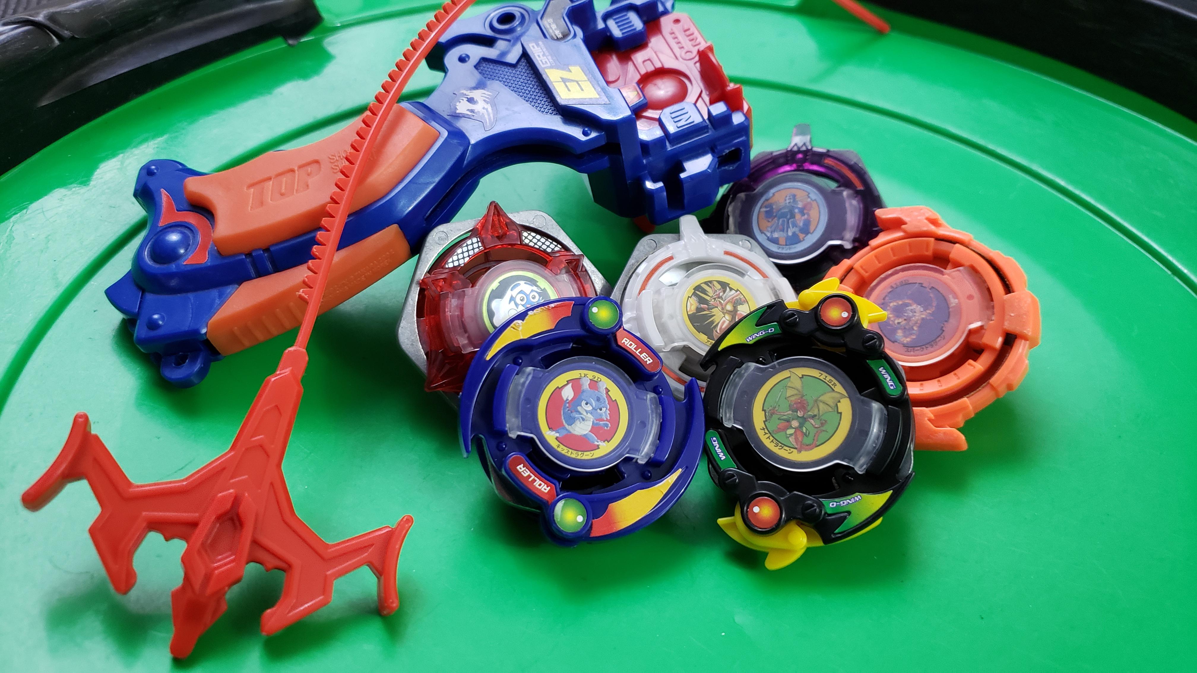 Several colourful Beyblade spinning tops and a blue-and-orange launcher arranged inside a bright green plastic arena.