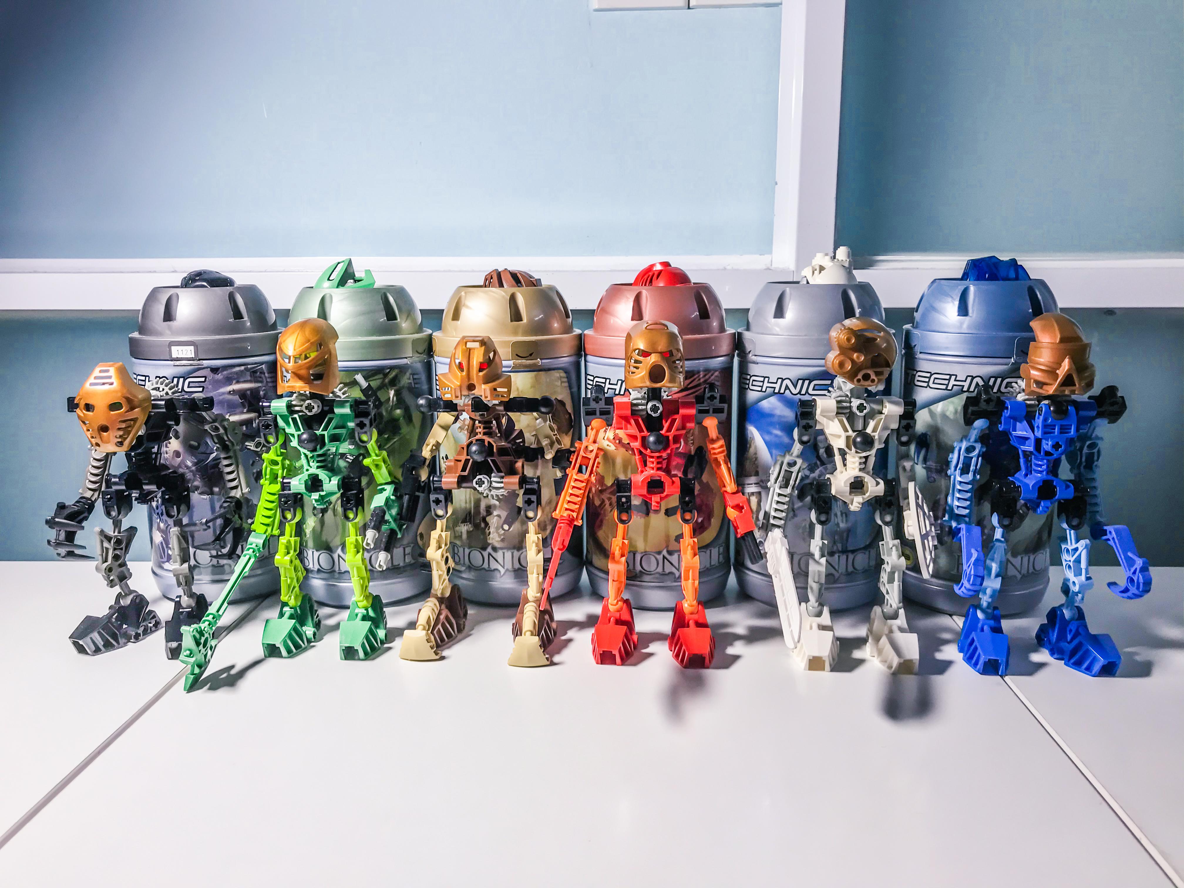 Six early Lego Bionicle figures in different colours standing in front of their cylindrical Technic-branded storage canisters.