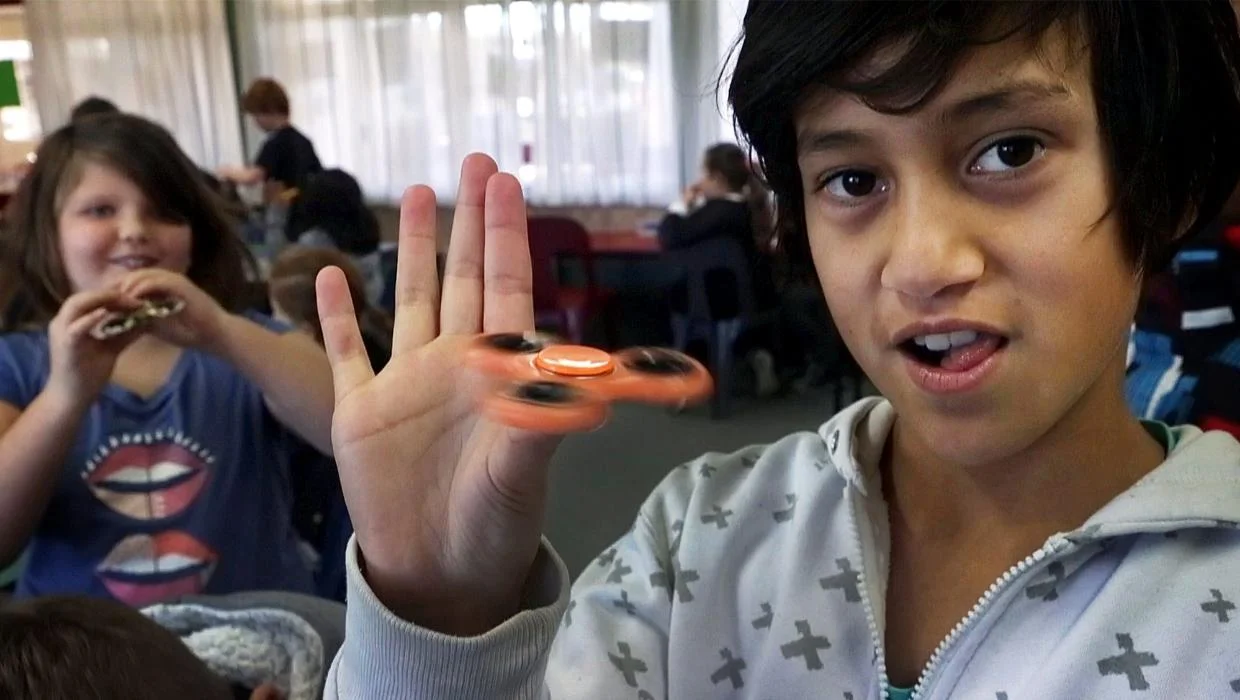 Child holding a spinning orange fidget spinner in front of an open hand while another child uses one in the background.