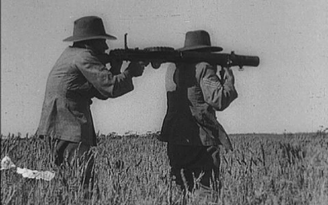 Black-and-white photograph of two men in wide-brimmed hats operating a machine gun in a field.