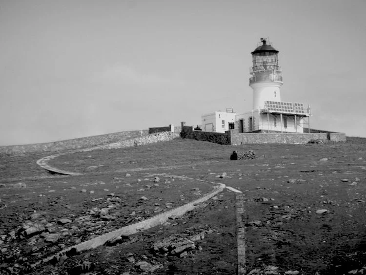 Black-and-white photograph of a solitary lighthouse and adjoining buildings on a barren rocky island.