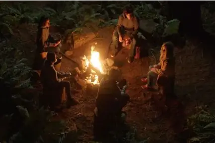 A group of young people seated in a circle around a campfire in a dark forest clearing.