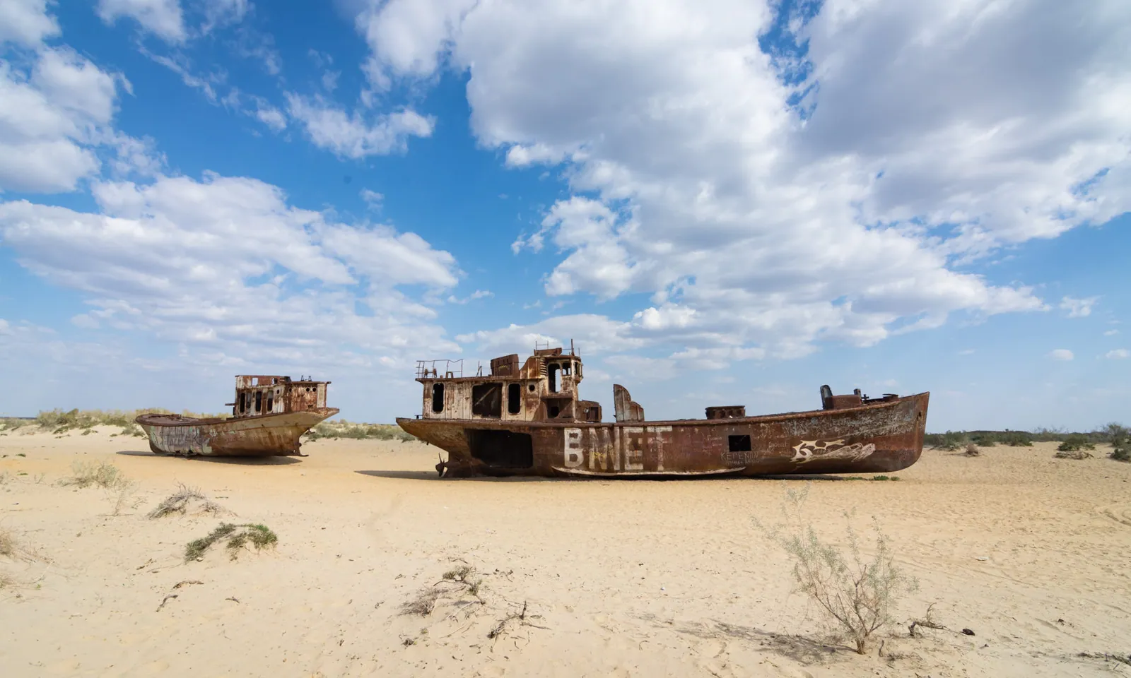 Two large rusting shipwrecks stranded on the sandy former bed of the Aral Sea beneath a broad cloudy sky.