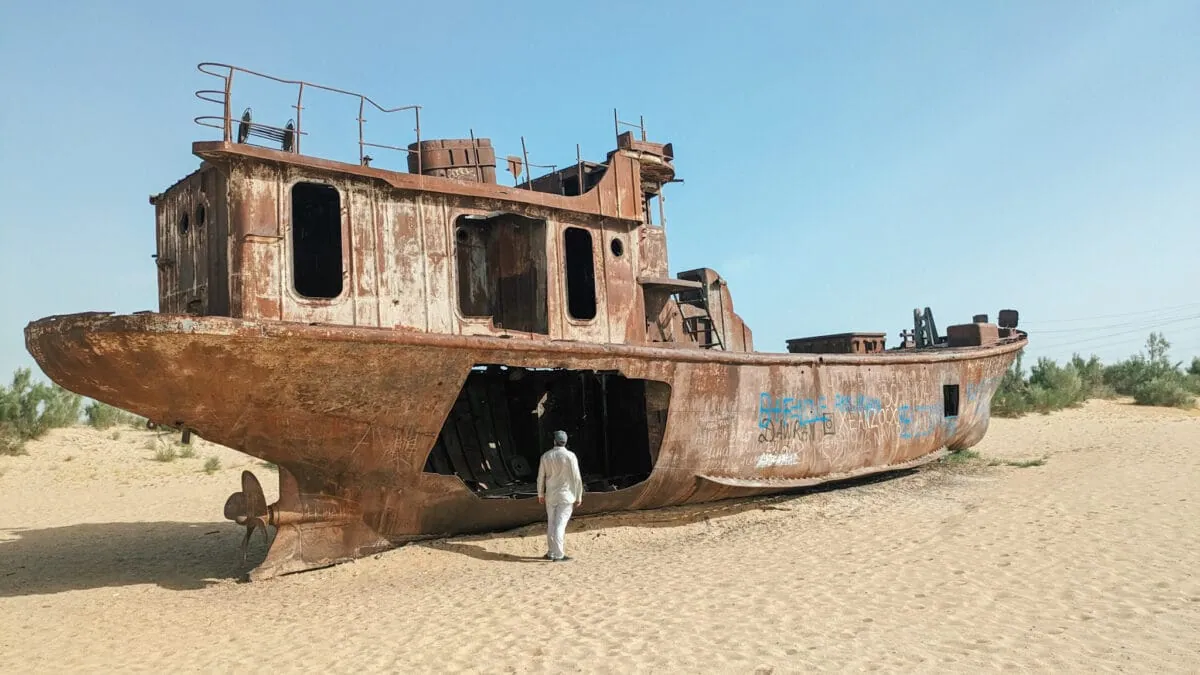 A person stands beside the corroded hull of a large shipwreck stranded in sandy desert.