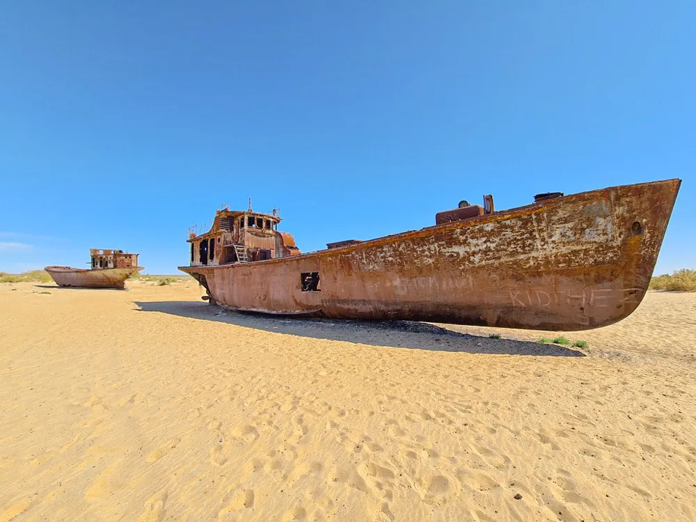 Two rusting shipwrecks rest on pale sand beneath a clear blue sky.