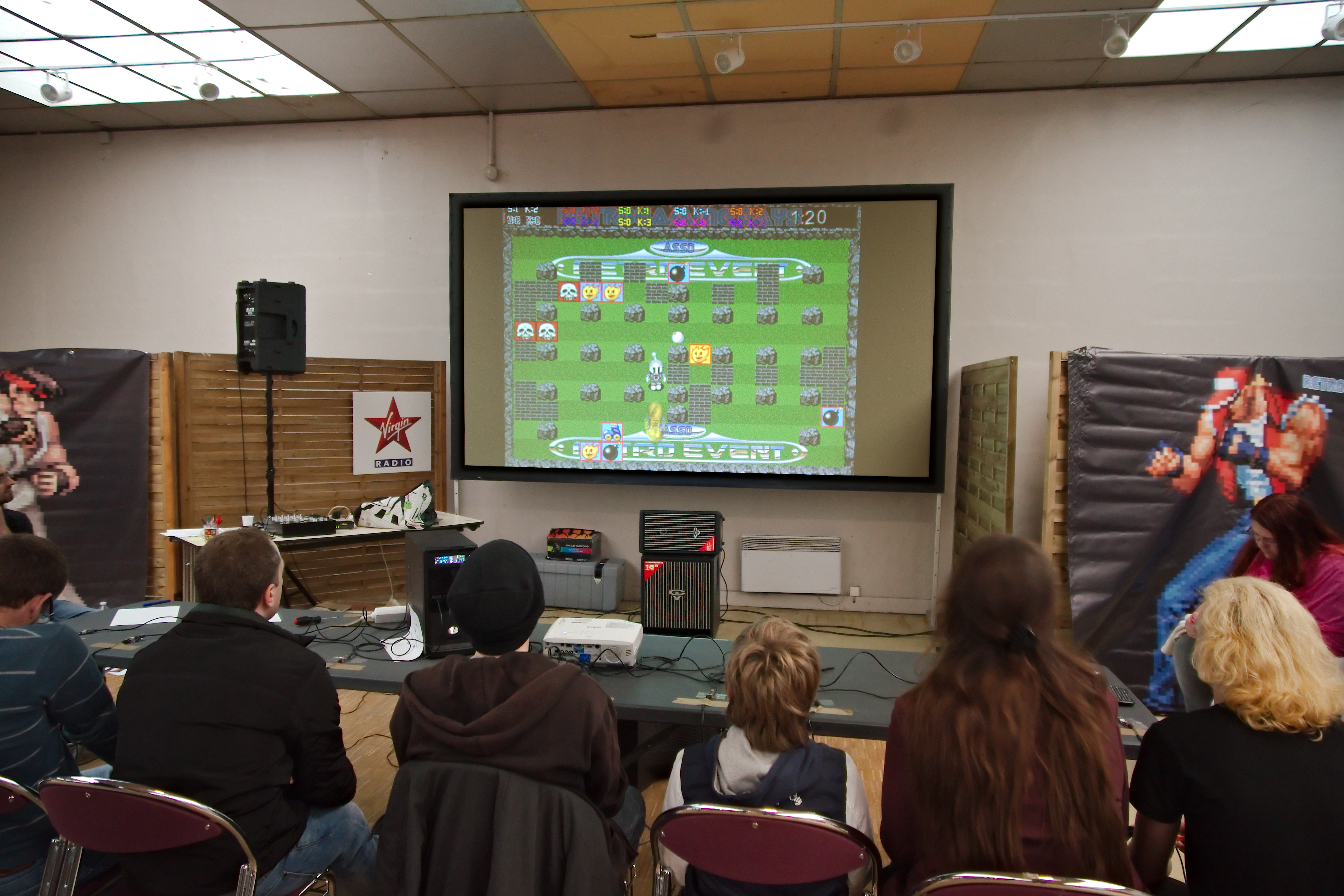 Audience watching a projected multiplayer Bomberman match at an indoor retro-gaming event.
