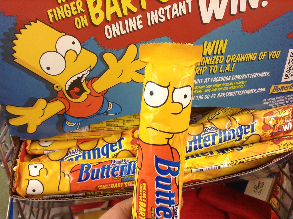 Store display of Butterfinger bars in Bart Simpson wrappers beneath a colourful promotional sign.