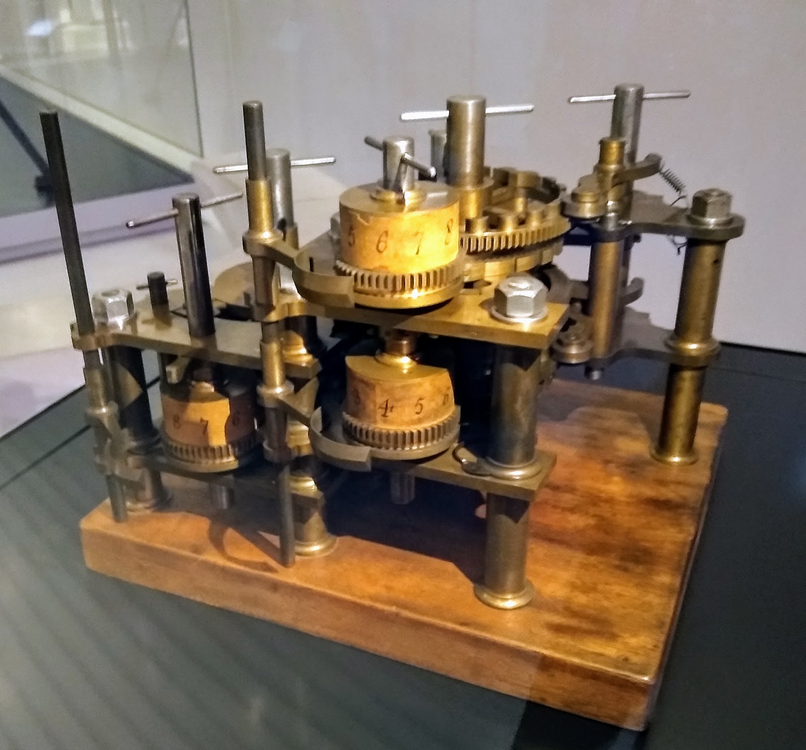 Small brass Difference Engine mechanism with numbered drums, gears and upright control rods on a wooden base.