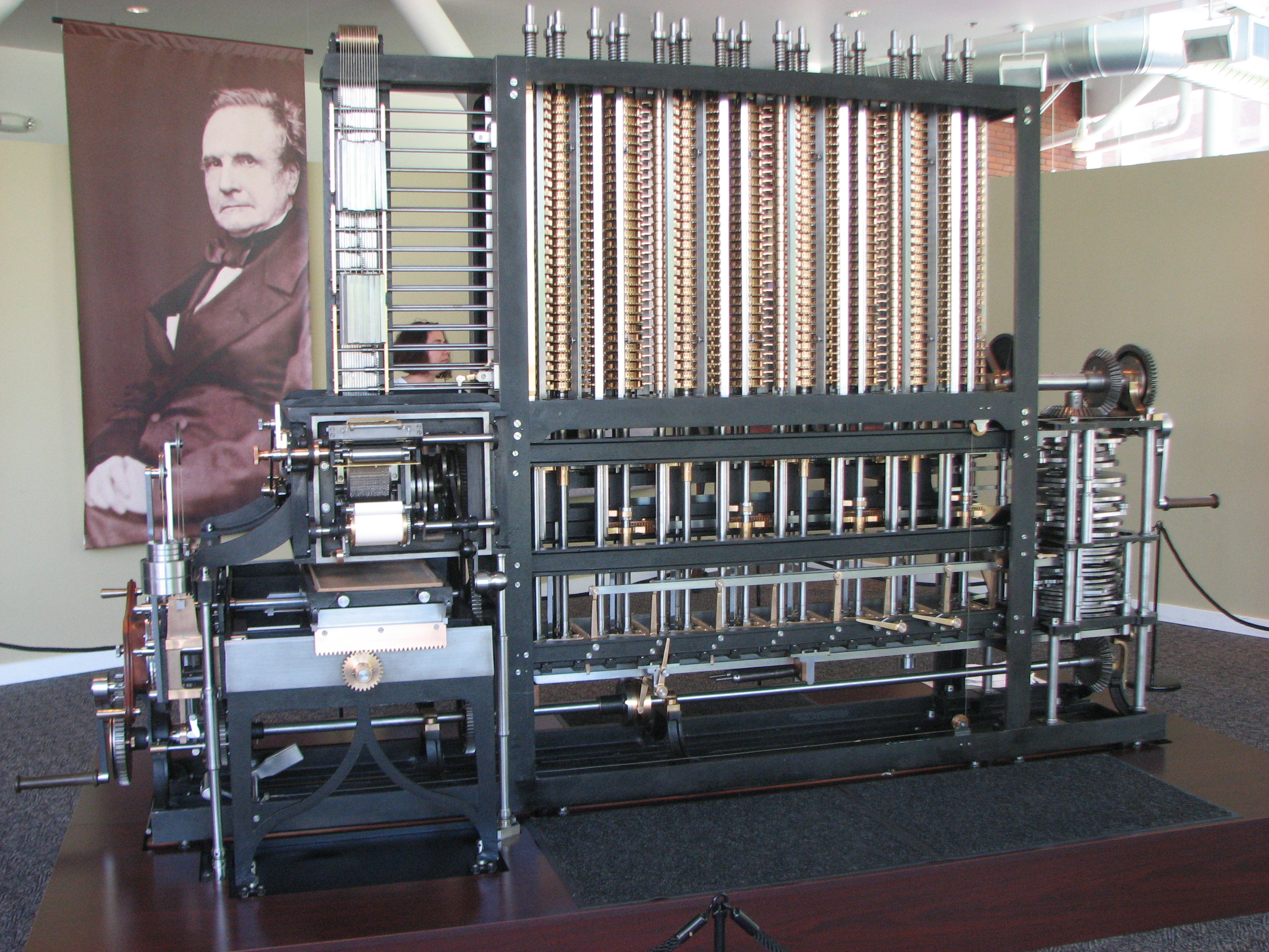 Large working Difference Engine reconstruction displayed beside a portrait banner of Charles Babbage.