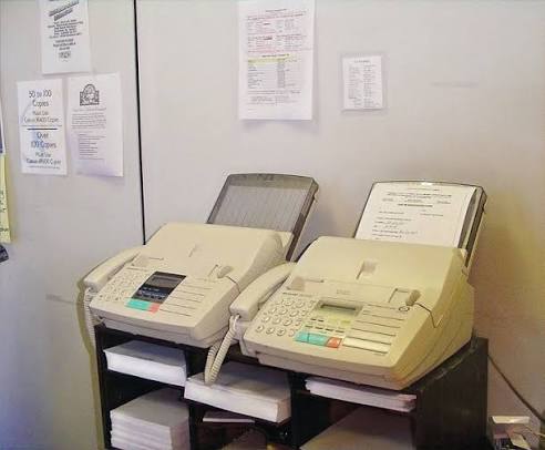 Two beige office fax machines standing side by side on paper-filled cabinets.