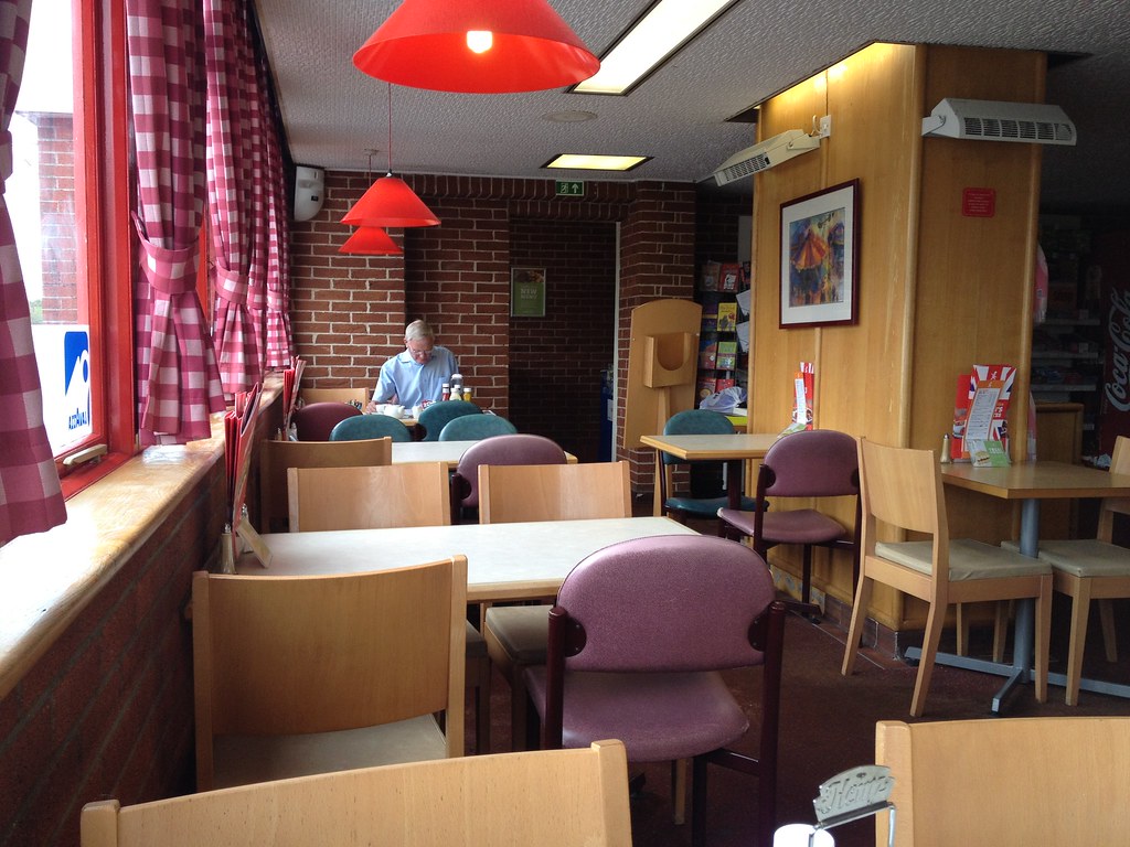 Interior of a Little Chef restaurant with wooden tables, purple chairs, red lamps and checked curtains.
