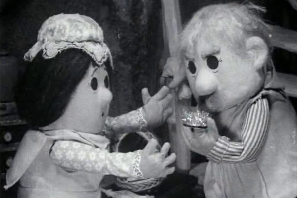 Black-and-white still of Mrs Pogle offering a basket while Mr Pogle drinks from a small cup.