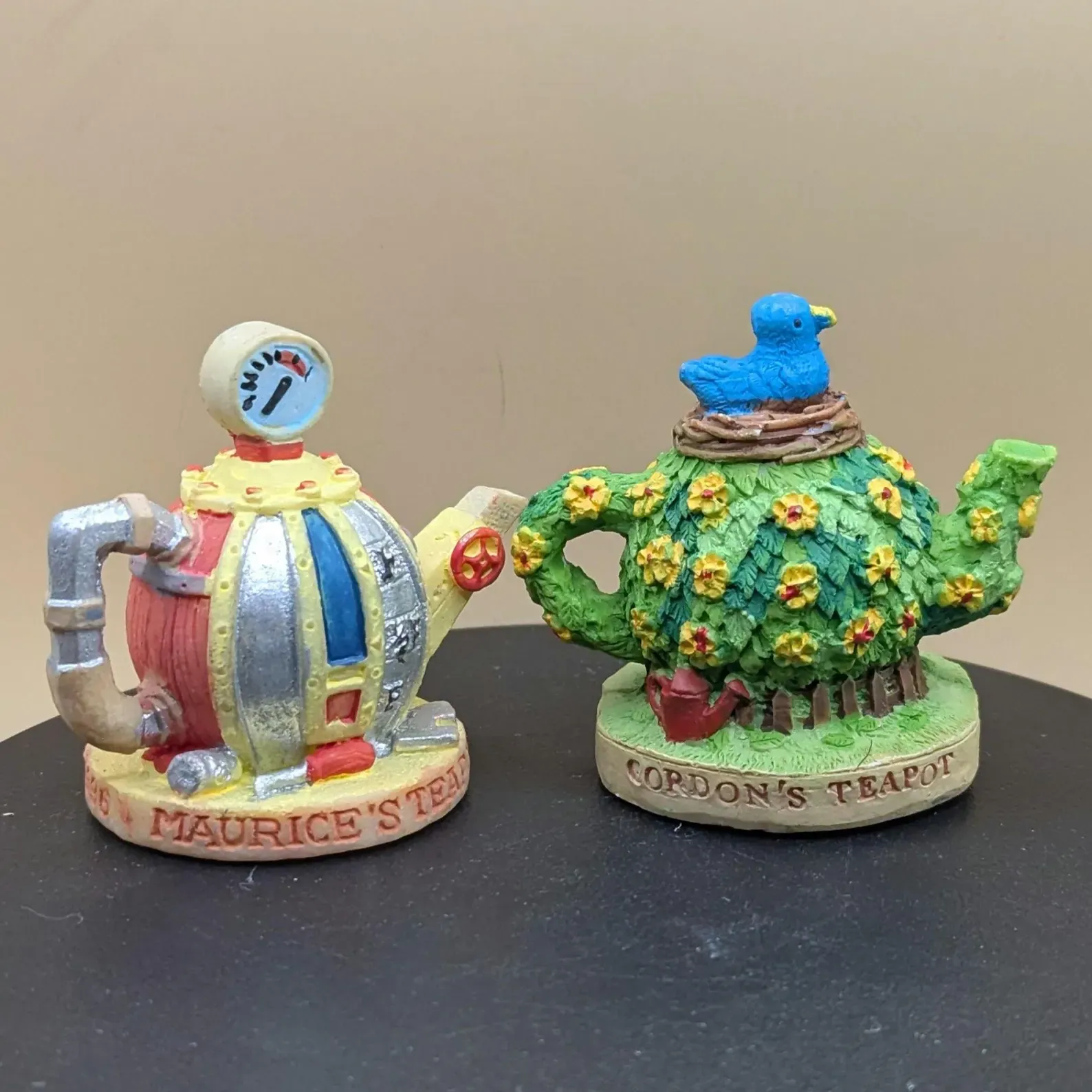 Two Tetley Tea Folk miniature ornamental teapots: Maurice's Teapot shaped like a colourful machine with pipes and a gauge, and Gordon's Teapot covered in green leaves and yellow flowers with a blue bird on top.