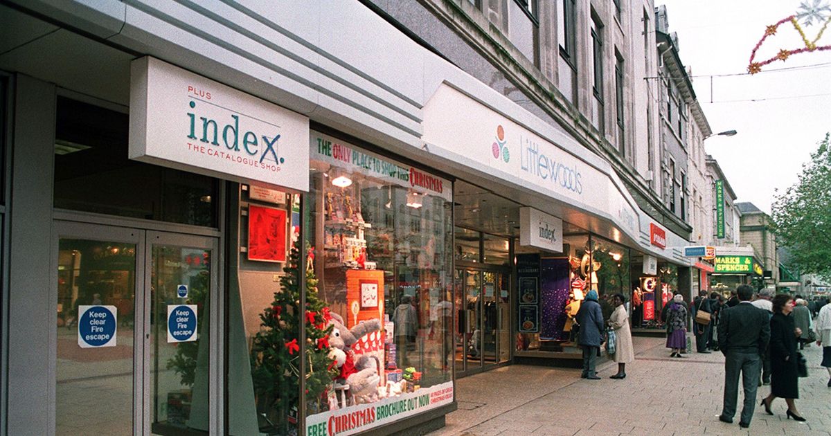 Street view of an Index The Catalogue Shop beside a Littlewoods storefront, with Christmas window displays and pedestrians on the pavement.