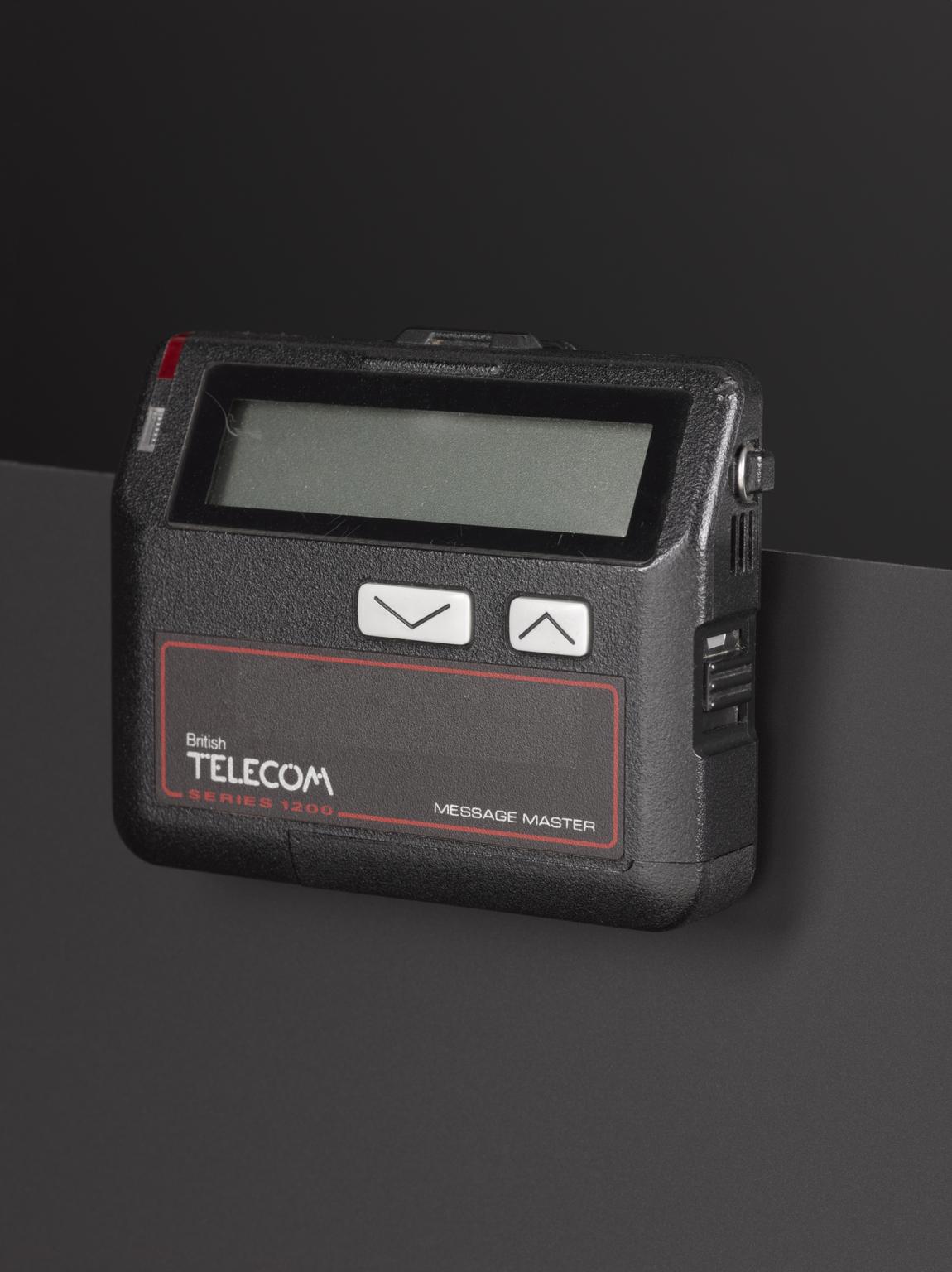 Black British Telecom Series 1200 Message Master pager standing on a dark surface, with a blank rectangular screen, two arrow buttons and red-outlined front label.