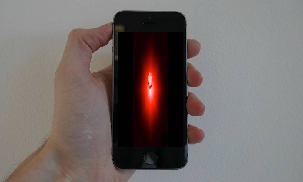 A hand holds a black touchscreen phone against a pale wall; its screen shows a small faceted red gemstone floating in a red glow on a black background.