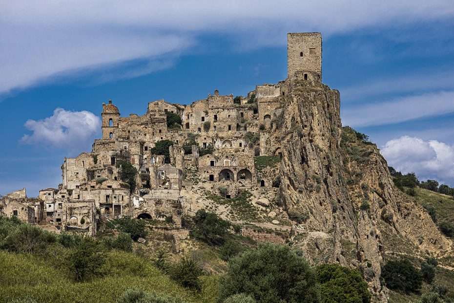 Wide view of a hilltop stone settlement with a square tower, crumbling buildings, a rocky cliff, green shrubs, and a blue sky with clouds.