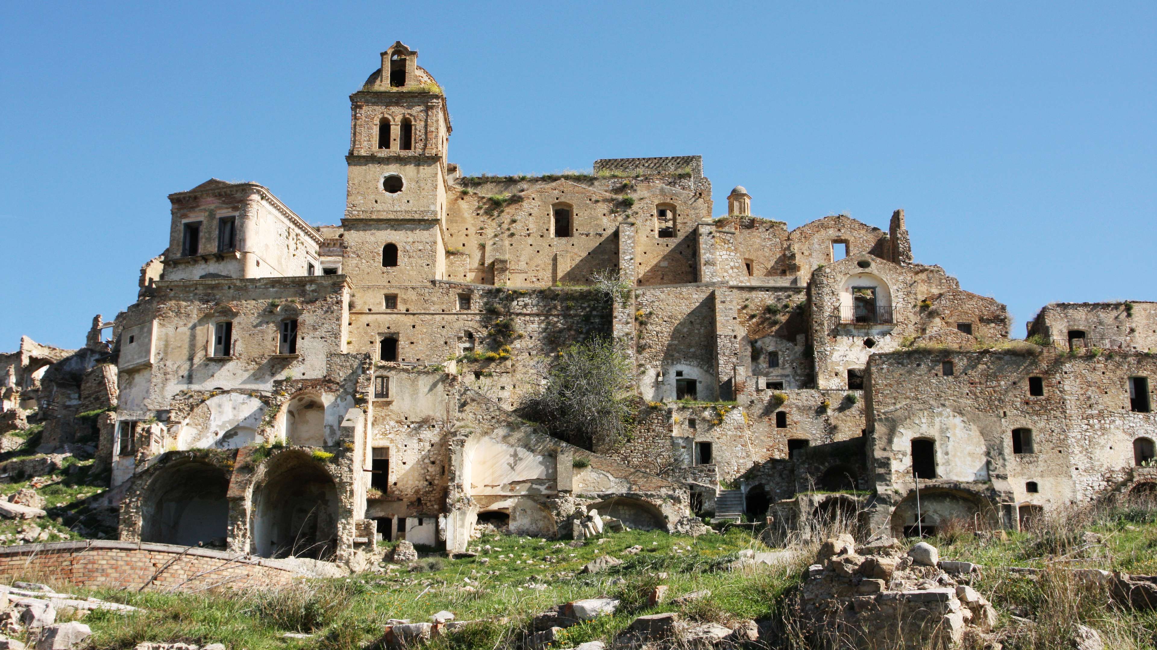 Ruined stone church and bell tower above stacked abandoned buildings with arched openings, broken walls, and grass-covered rubble under a clear blue sky.