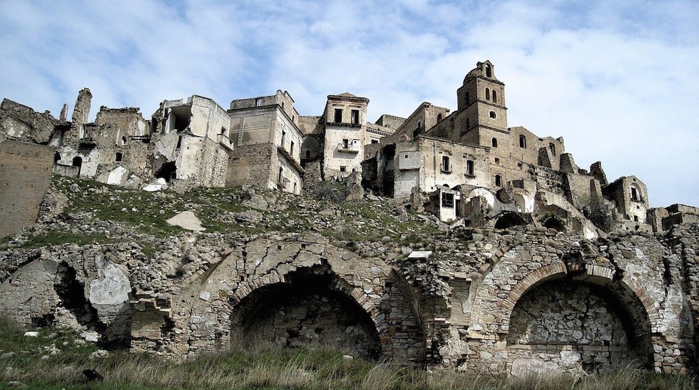 A row of damaged stone buildings and a bell tower rise behind three large collapsed stone arches on a rocky slope.