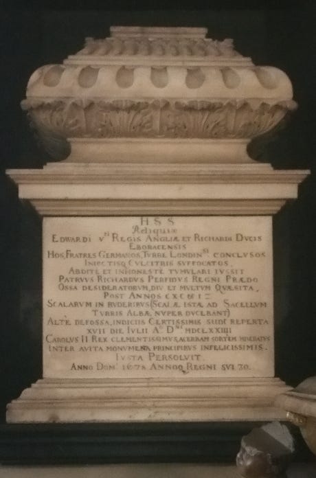 Close view of a pale carved stone urn on a stepped base, with an inset plaque bearing multiple lines of Latin text against a dark background.