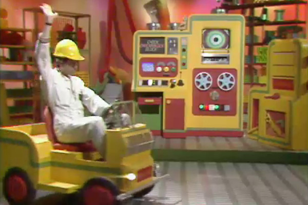 A person in a white outfit and yellow hard hat drives a small yellow vehicle in front of a large yellow-and-green control machine with a screen, buttons, lights and two reel-like wheels.