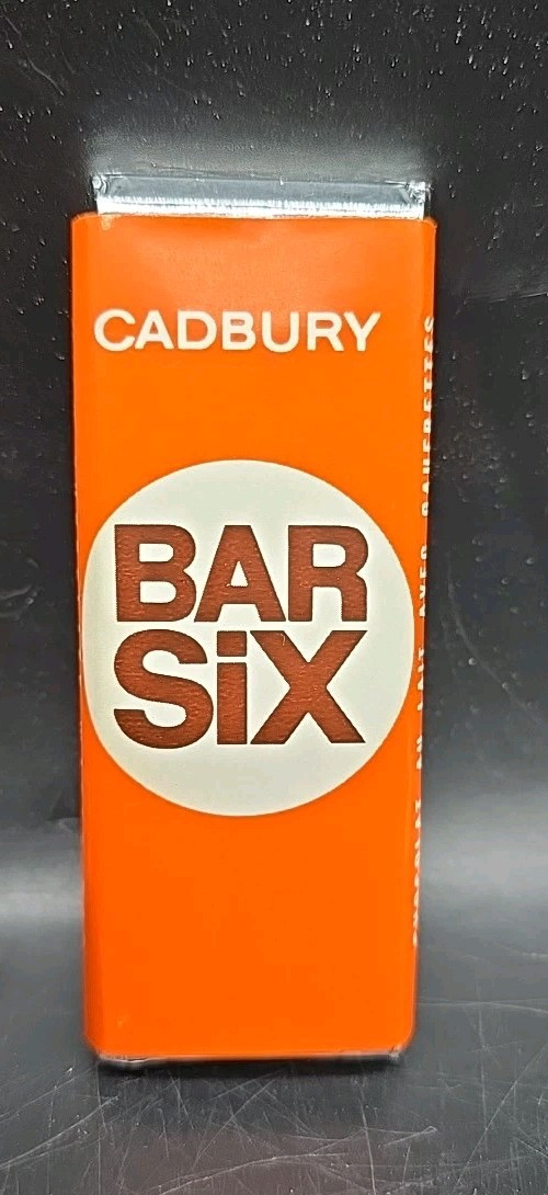 Front view of an orange Cadbury Bar Six chocolate-wafer-bar wrapper standing upright against a dark speckled background, with exposed silver foil at the top and a large white circle containing brown “BAR SIX” lettering.