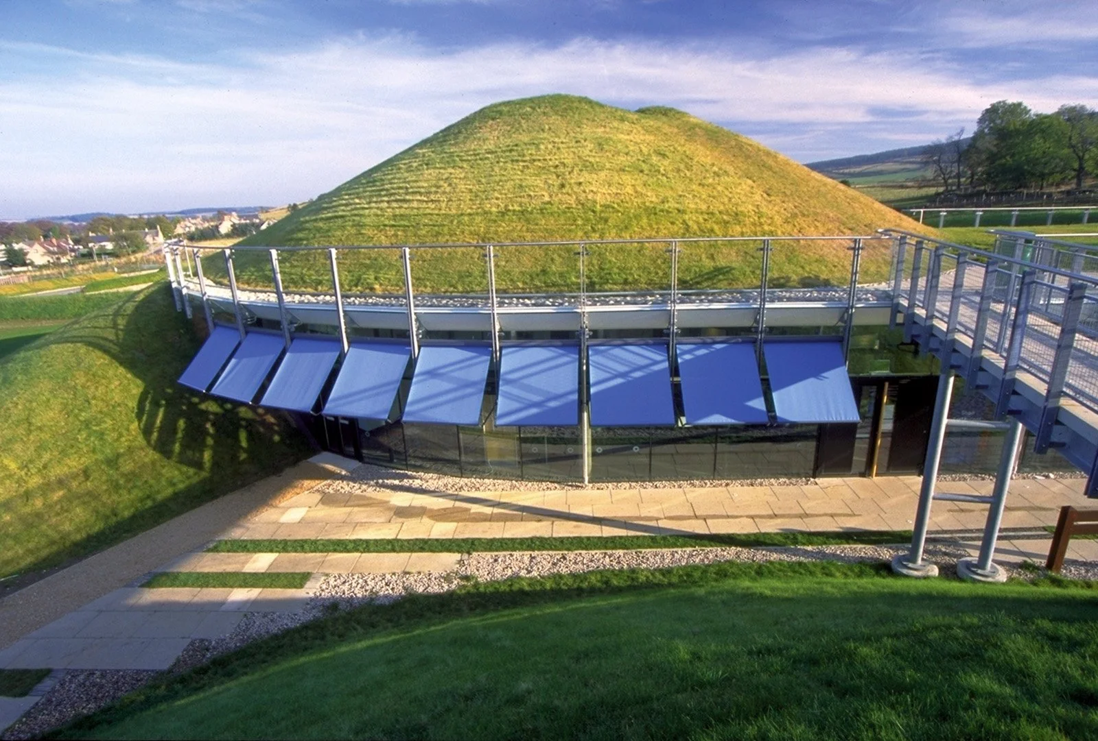 Elevated view of a large grass-covered mound above a glass-fronted building with blue awnings, metal railings, paved paths and surrounding fields under a cloudy sky.