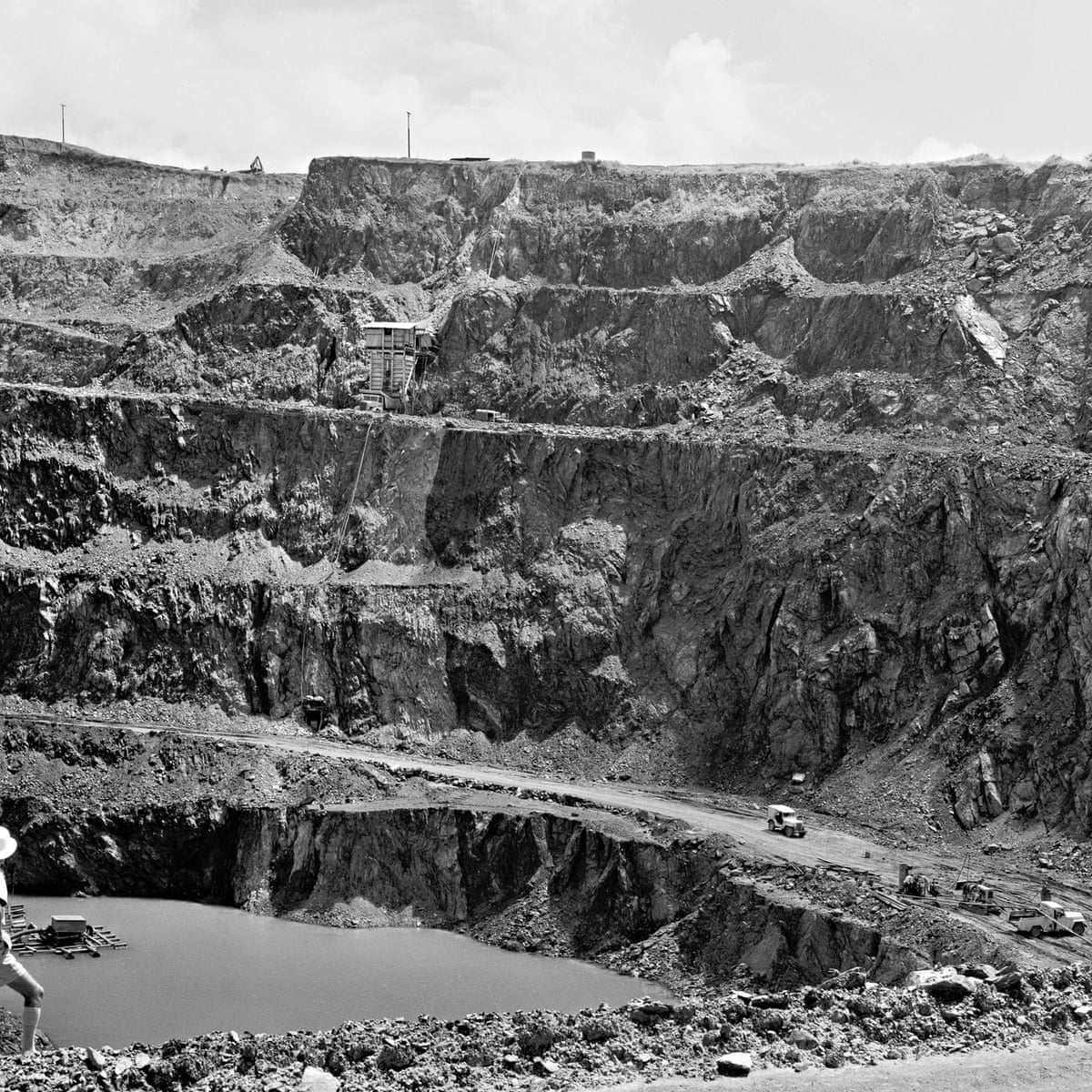 Black-and-white wide view into a terraced open-cut mine, with steep rock benches, a haul road, small vehicles and equipment, and a pool of water at the bottom left.
