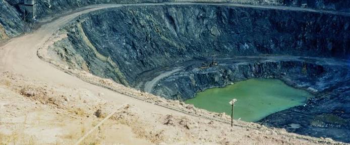 View down into a stepped rock open pit containing a small pool of opaque green water, with a pale dirt haul road curving around the rim and benches.