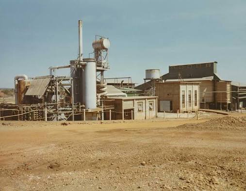 Wide view of an industrial processing plant with metal tanks, pipes, conveyors, a tall smokestack and low rectangular buildings behind a broad dirt foreground under a pale blue sky.