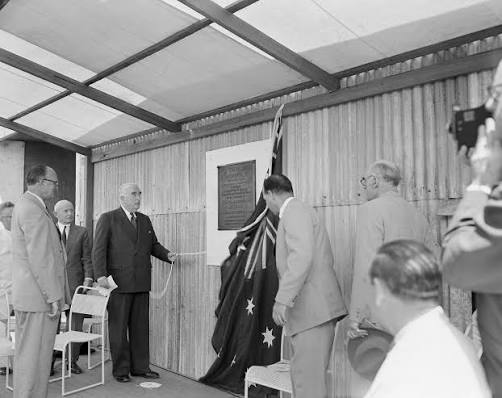 Black-and-white photograph of several suited men at a small outdoor ceremony beside a corrugated wall, as an Australian flag is pulled aside to reveal a wall plaque; seated attendees and a camera operator are visible.