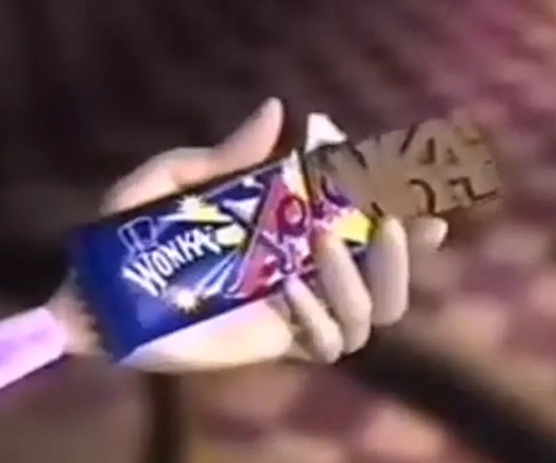 Close-up of a hand holding a partly unwrapped Wonka chocolate bar. The blue and pink wrapper shows the white Wonka logo, and raised lettering is visible on the exposed chocolate.