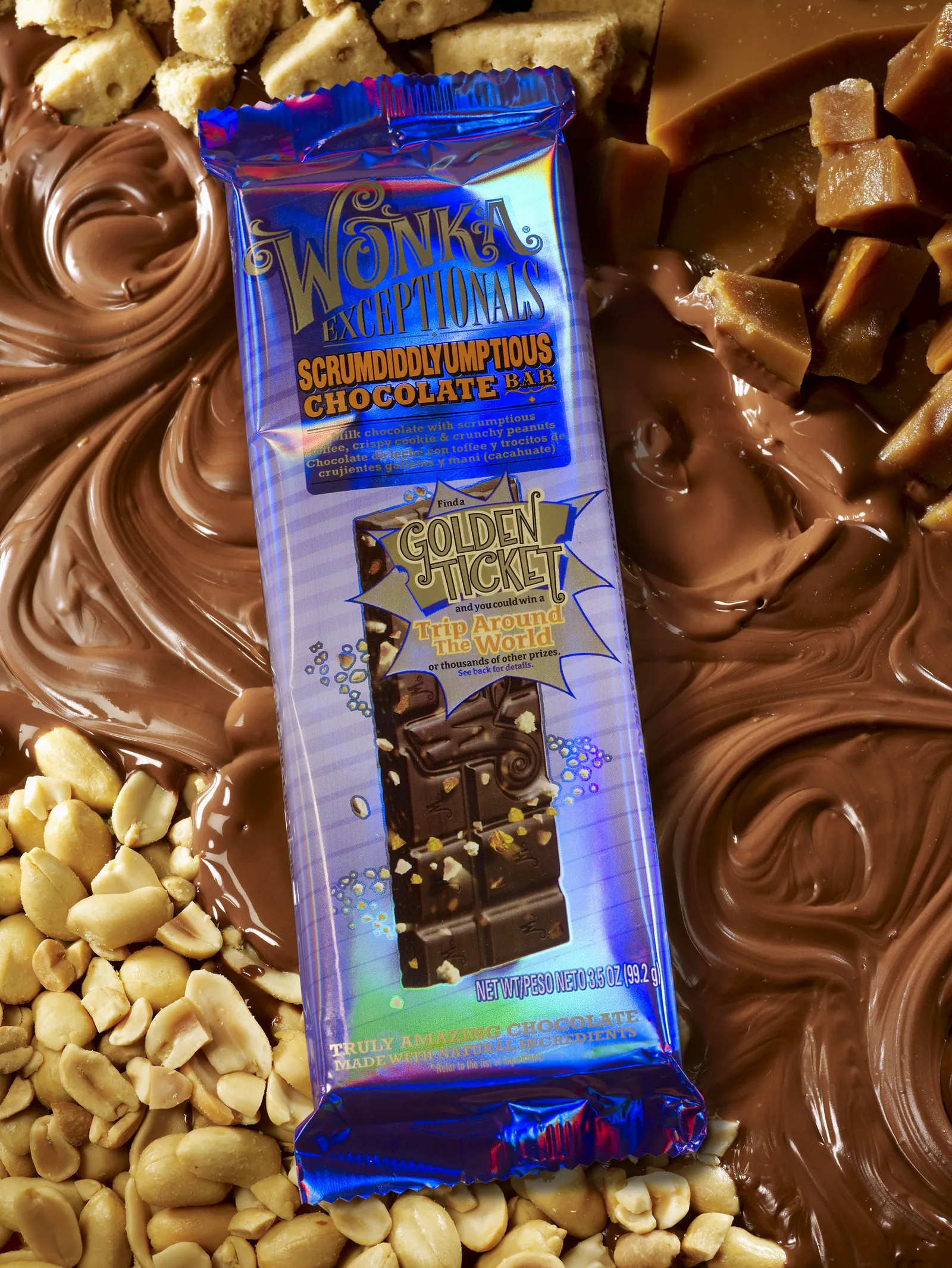 Close-up of a blue-and-purple Wonka Exceptionals Scrumdiddlyumptious Chocolate Bar wrapper lying on swirled milk chocolate, with peanuts, cookie pieces, and caramel pieces around it. The wrapper advertises a golden-ticket prize promotion and shows pieces of chocolate with nuts.