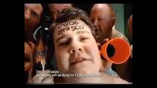 Low-resolution close-up commercial still of a young man's face marked with dark writing on his forehead, surrounded by men and two orange megaphone openings; small white text appears along the bottom.