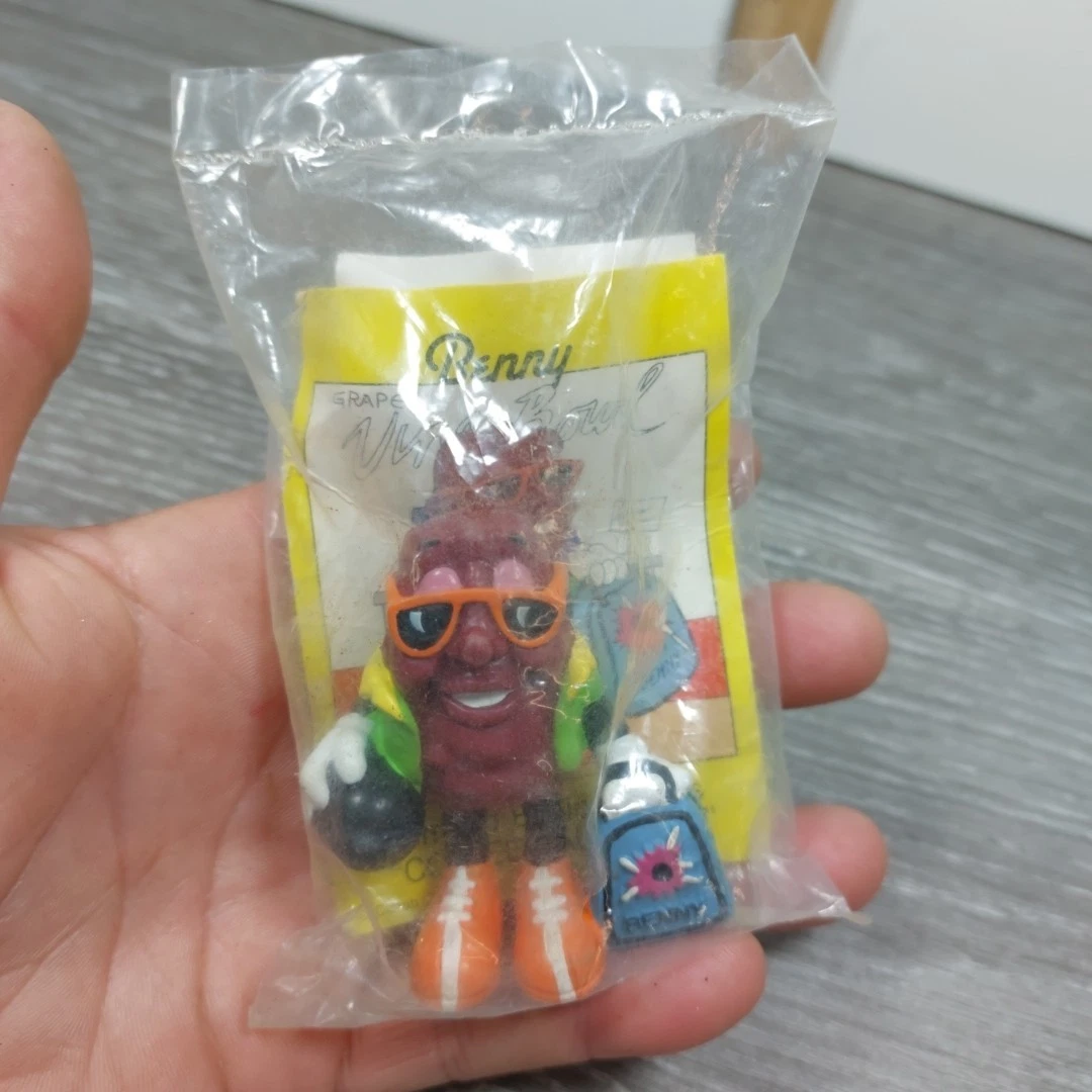 A hand holds a sealed clear plastic bag containing a burgundy California Raisins figurine wearing orange sunglasses and orange shoes, a small blue accessory, and a yellow-backed card.