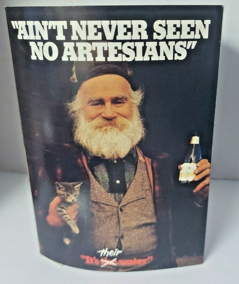 Photograph of a printed Olympia advertisement or poster. Large white text at top reads “AIN’T NEVER SEEN NO ARTESIANS.” Below, a bearded man in a dark cap holds a kitten in one hand and a beer bottle in the other. Small text and a red slogan run along the bottom edge.