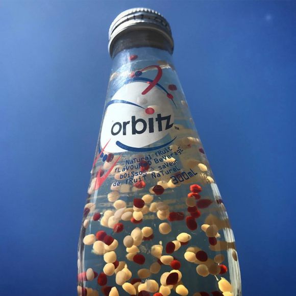 Low-angle close view of an Orbitz bottle against a blue sky, with a white cap, a white Orbitz label, clear liquid, and red and pale yellow spheres inside.