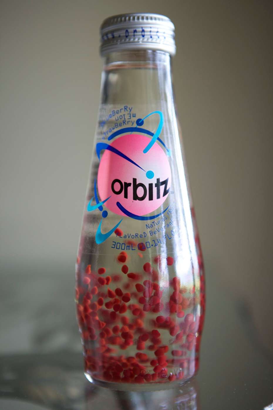 Front-facing clear Orbitz bottle standing on a reflective surface, with a white cap, pink Orbitz logo, and numerous red spheres suspended in the liquid against a softly blurred gray background.