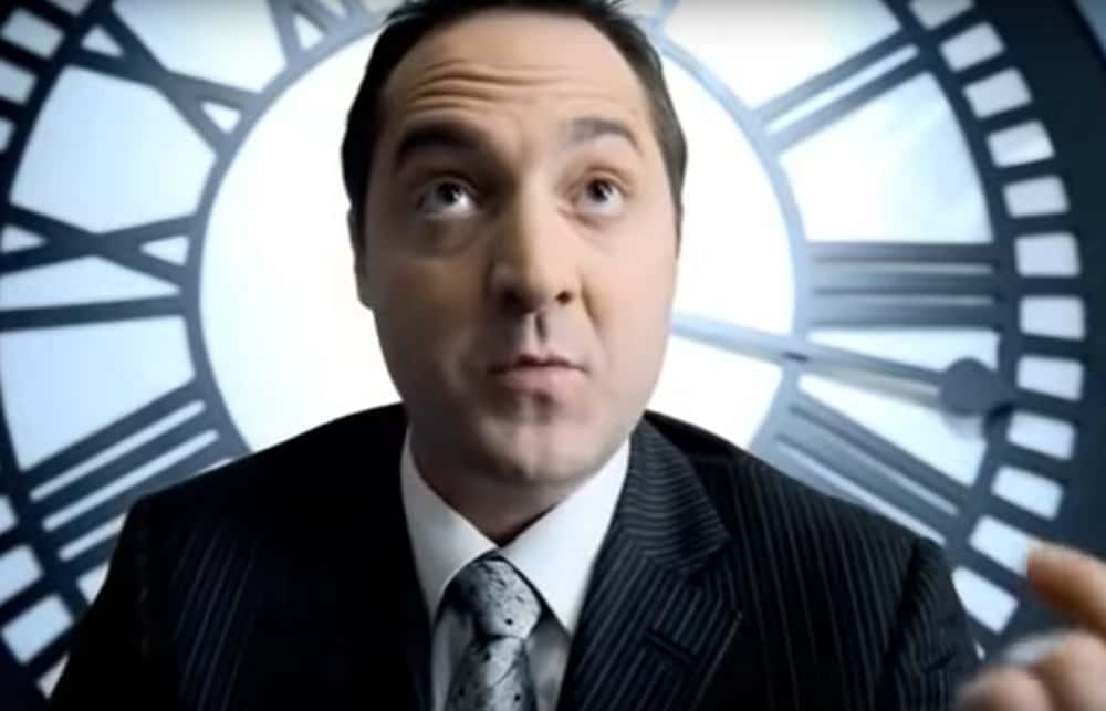 A man in a dark pinstripe suit and patterned tie looks upward in front of a large clock face; part of a hand is visible at the right edge.