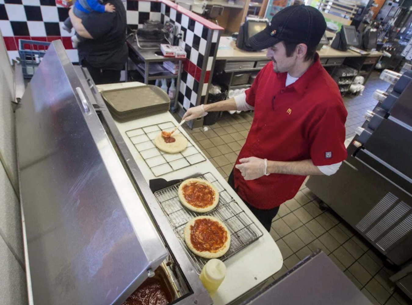 Overhead view inside a McDonald's kitchen: a man in a red McDonald's uniform spreads tomato sauce on a round pizza base while two sauced bases sit on wire racks beside a large metal oven or preparation unit.