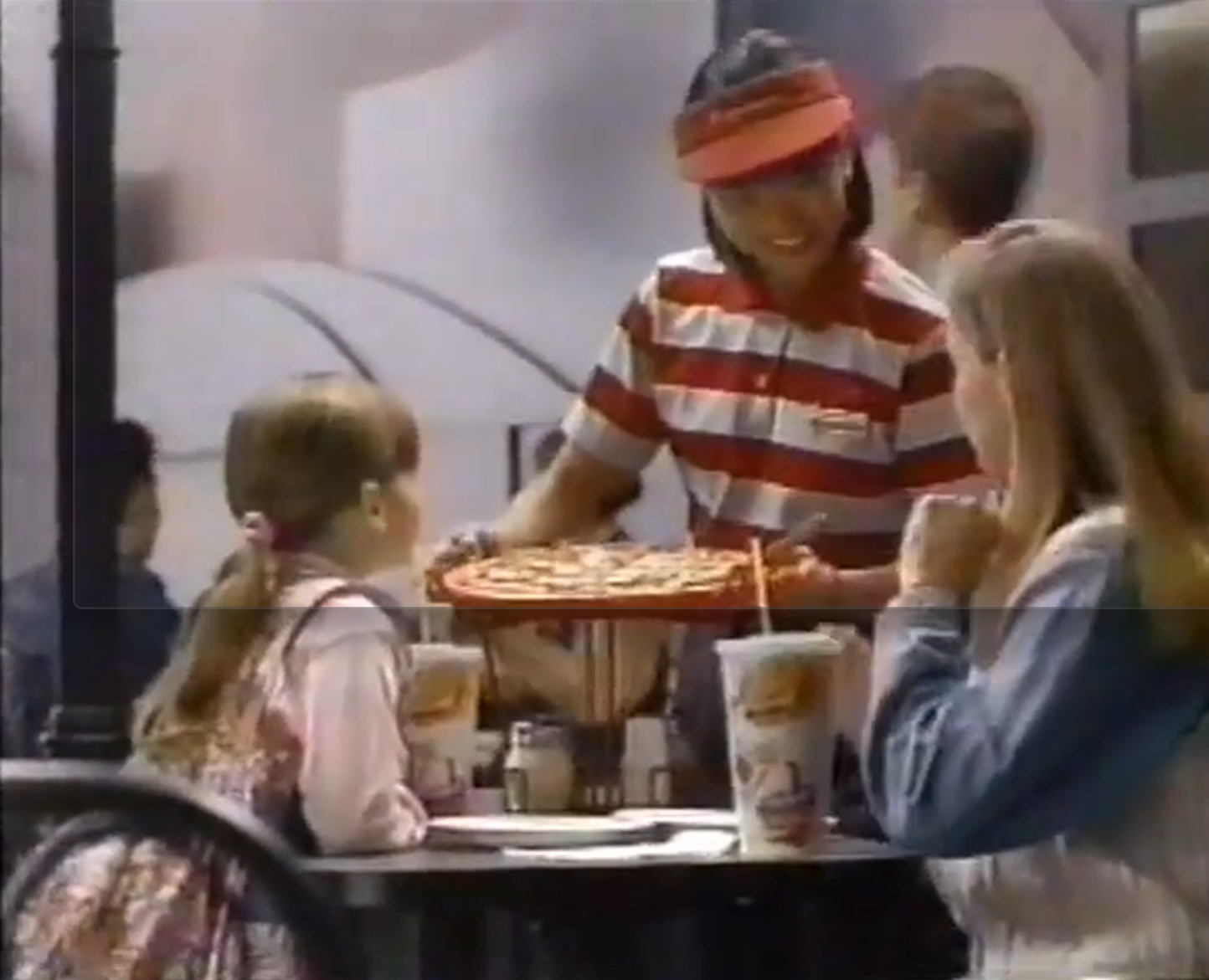 Soft-focus image of a McDonald’s employee in a red-and-white striped shirt and visor serving a pizza on a tray to adults and children seated at a restaurant table with paper drink cups.
