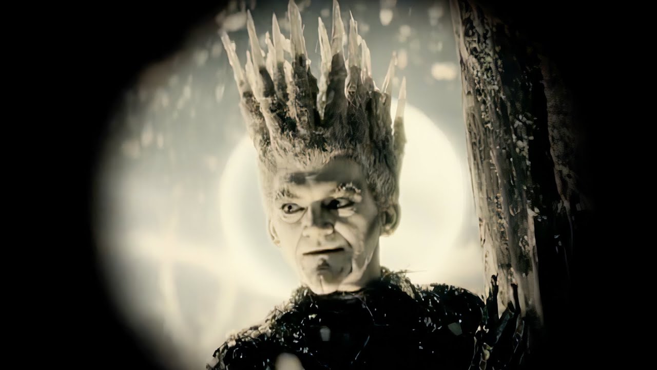 Close-up of a pale stern-faced figure wearing a tall jagged crown-like headpiece, framed between dark tree trunks with a bright circular light behind the head.