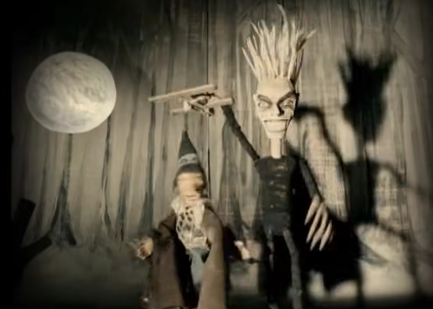 Two stylized figures stand before vertical tree trunks beneath a large full moon: a tall pale-faced figure with spiked hair and long fingers holds a cross-shaped object, while a smaller bearded figure stands beside it.