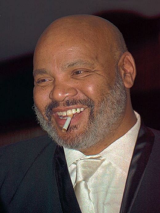 Close-up color portrait of a smiling bald man with a gray beard wearing a dark tuxedo jacket, white shirt, and pale tie, with a cigarette held between his teeth.