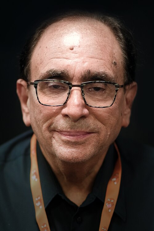 Close-up color portrait of a middle-aged man wearing rectangular eyeglasses, a dark collared shirt, and an orange lanyard against a black background.