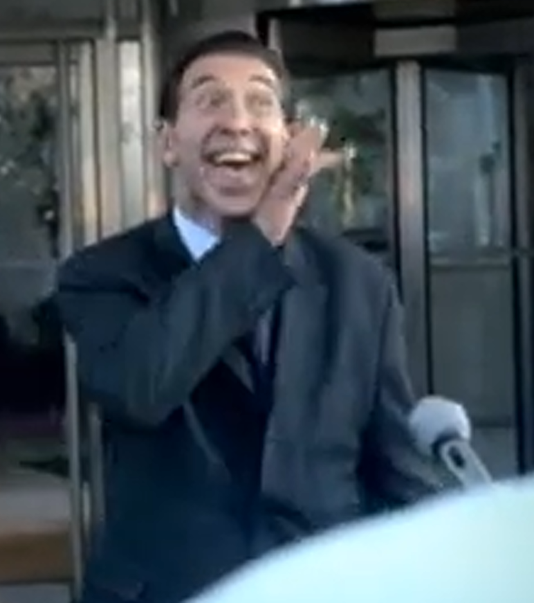 Low-resolution frame of a smiling man in a dark suit outside glass doors, raising one hand beside his face; a microphone and a pale foreground object appear at lower right.