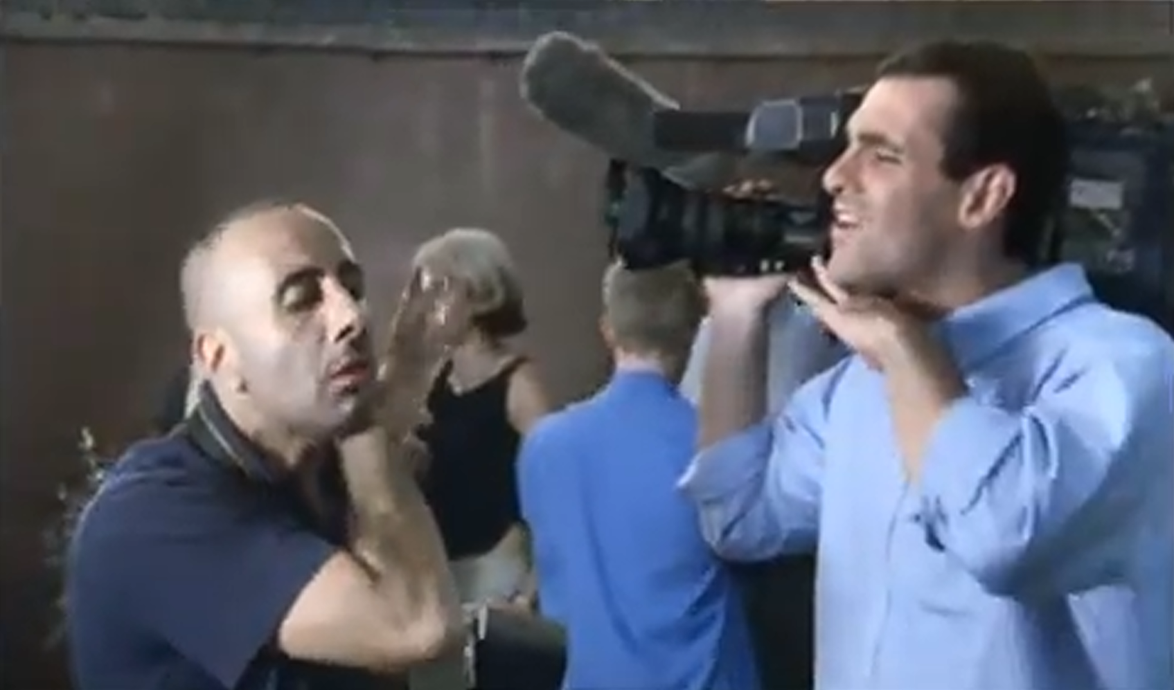 Low-resolution video frame showing a bald man in a dark shirt beside a man in a pale blue shirt who is holding both hands near his neck; a large video camera and several people appear behind them.