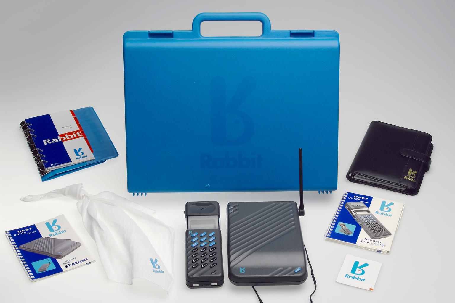 Studio photograph on a pale background of a blue Rabbit-branded hard carrying case, a dark cordless handset, a ribbed base unit with an upright antenna, two user guides, a black leather case, a white drawstring bag and small Rabbit-branded papers.